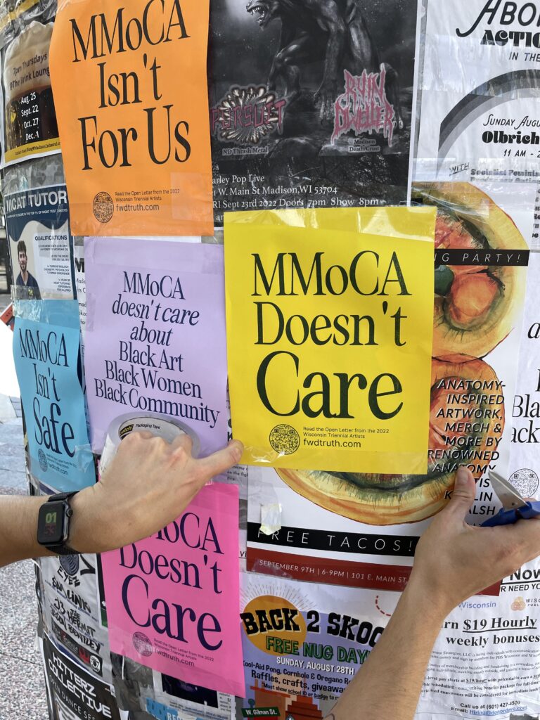 Hands post a series of awareness raising posters on a public signboard. Posters read: "MMoCA Doen'st Care," "MMoCA Isn't For Us," and "MMoCA Doesn't Care Abotu Black Art, Black Artists, Black Community."